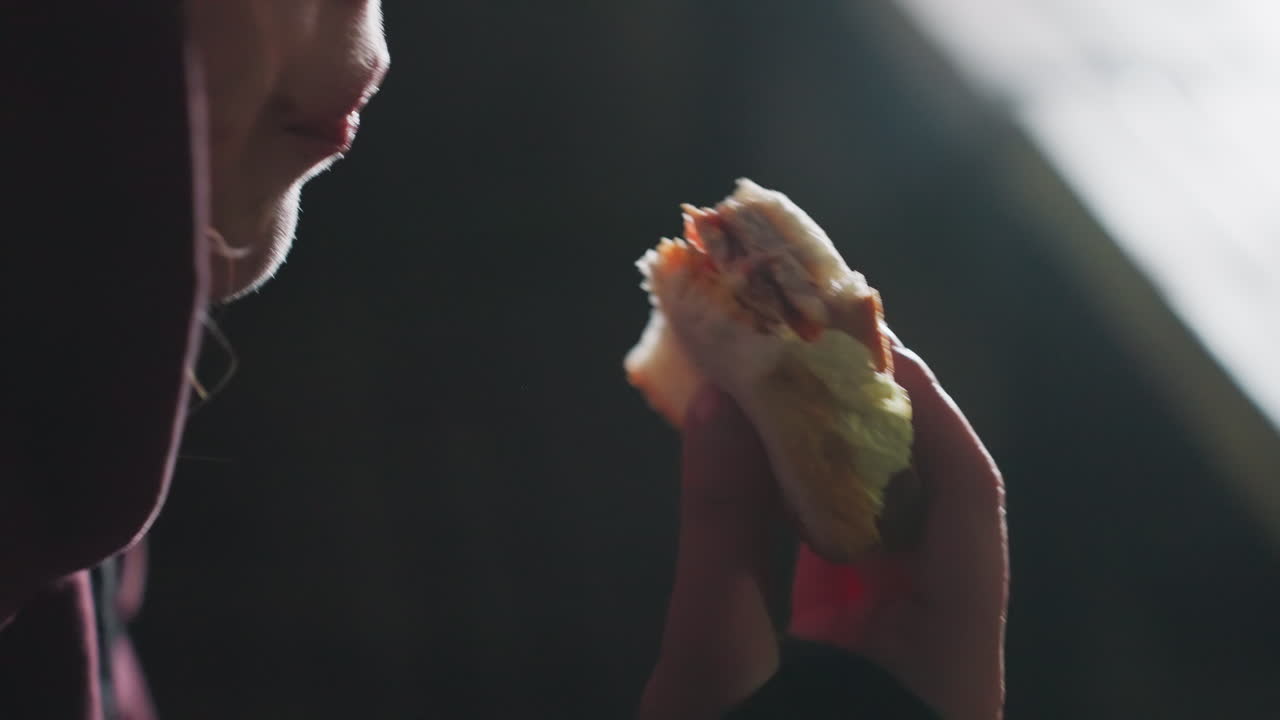 Close up side profile of hoodie athlete eating bread sandwich and sipping water from sports bottle at twilight outdoors under cool air post workout recovery moment glowing urban lights