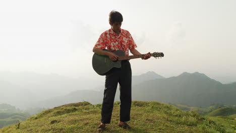 asian male musician enjoying strumming guitar and singing on mountain top