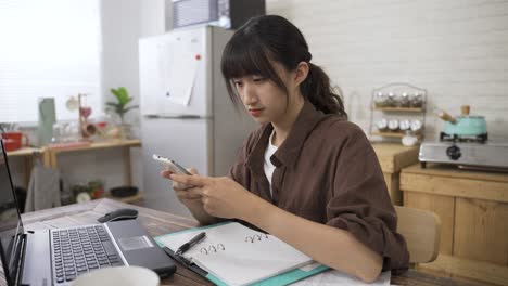 portrait of a serious asian businesswoman texting message for work on the phone with concentration while working from home in the dining room at daytime.