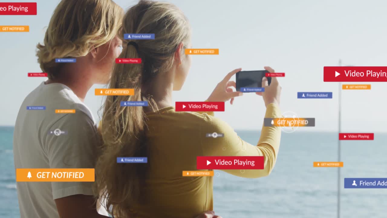 스마트 폰에서 바다의 사진을 찍는 백인 부부의 뒷면에 여러 개의 디지털 아이콘이 있습니다.