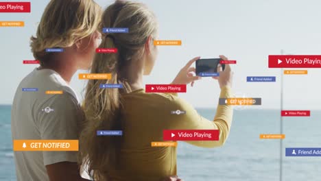 Mehrere-Digitale-Symbole-über-Der-Rückansicht-Eines-Kaukasischen-Paares,-Das-Mit-Dem-Smartphone-Das-Meer-Fotografiert