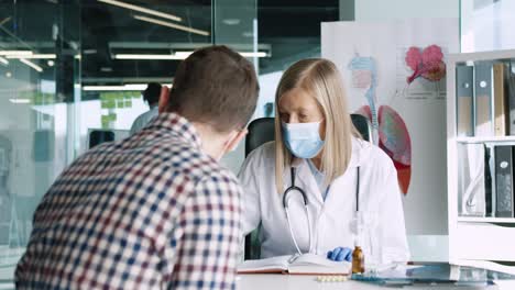 Rear-view-of-male-patient-sitting-at-desk-and-speaking-with-senior-female-doctor-in-medical-consultation