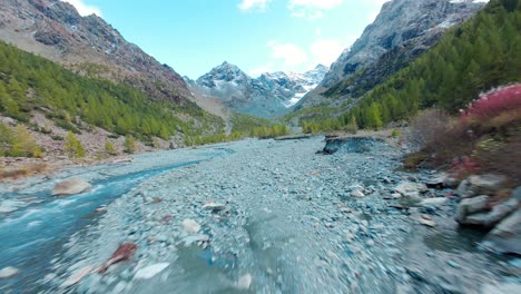 aerial fpv perspective of the valmalenco alps in italy, showcasing a serene river flowing through majestic mountains and lush greenery on a clear, sunny day.