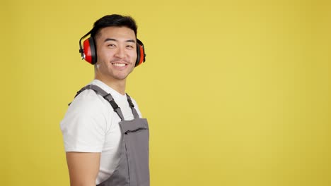 happy asian man in work uniform and ear protection smiling on yellow background