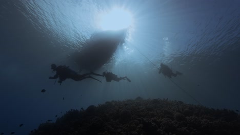 Toma-De-Luz-De-Fondo-De-Buceadores-Bajo-Un-Bote-De-Buceo-En-Aguas-Azules-Claras-Junto-A-Una-Silueta-De-Arrecife-De-Coral-Tropical,-Toma-De-Belleza-En-La-Isla-De-Tahití,-Polinesia-Francesa