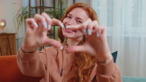 happy young caucasian woman sitting on couch makes symbol of love showing heart sign to camera