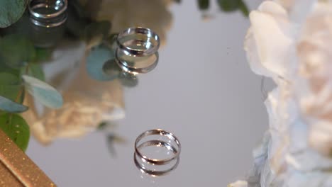 Three-wedding-rings-rolling-on-a-glass-table,-surrounded-with-white-flowers-and-wedding-details