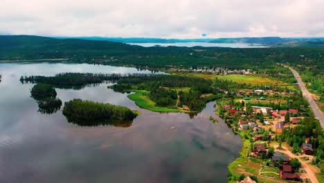 aerial drone view of a peaceful clear lake with cars driving next to a mountain town with cabins and vacation houses in a forest with cloudy hills in the background