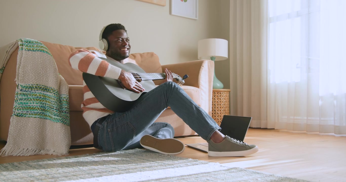 Homme afro-américain assis par terre chez lui avec un casque audio, suivant une piste sur son ordinateur portable.