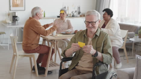 medium long pov of senior caucasian man with yellow sticky note saying fox on forehead, smiling, talking on camera, sitting in wheelchair in foreground of blurred people