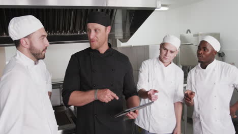 diverse male chef instructing group of trainee male chefs using tablet in kitchen, slow motion