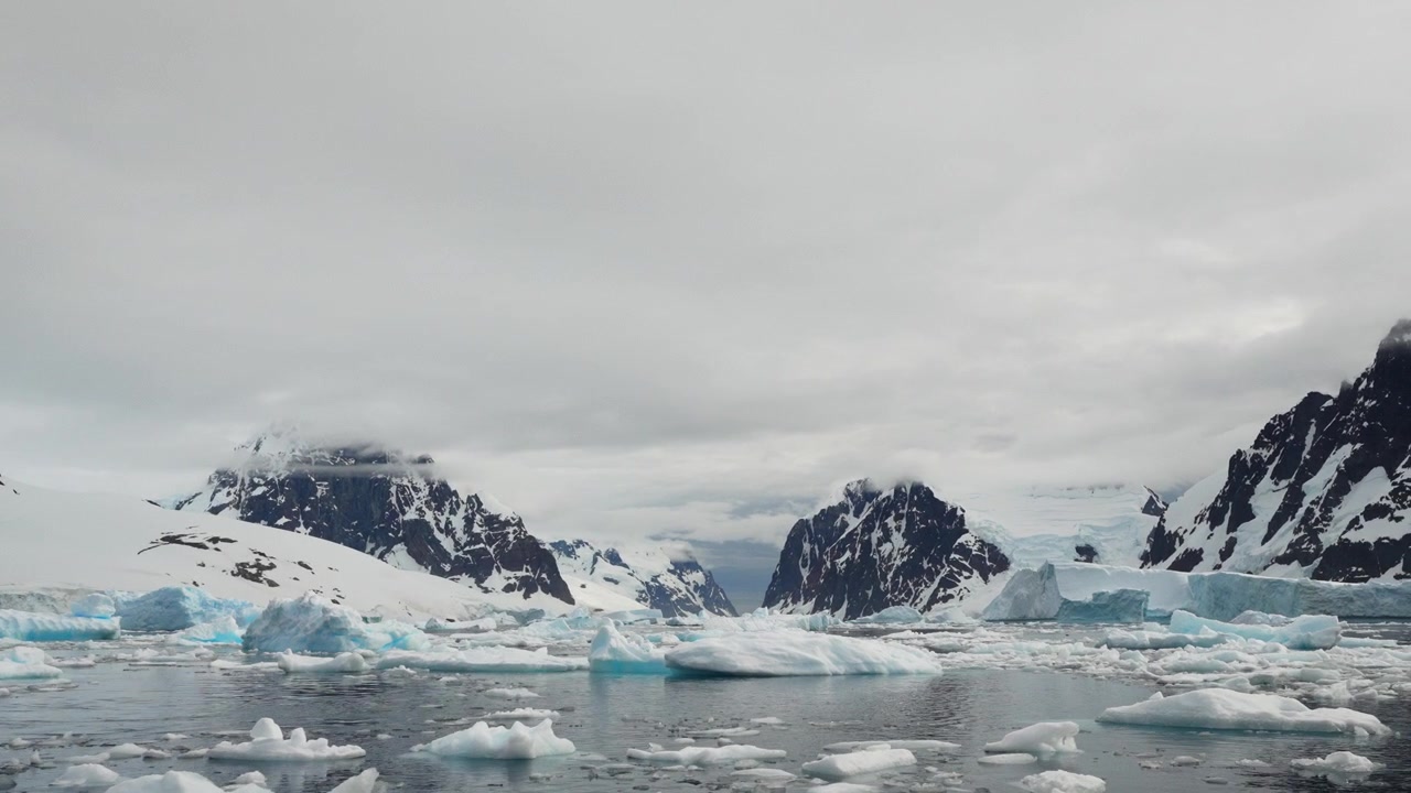 Oceano Antartico ghiacciato con iceberg e banchi di ghiaccio galleggianti, montagne rocciose coperte di neve sotto un cielo coperto sullo sfondo