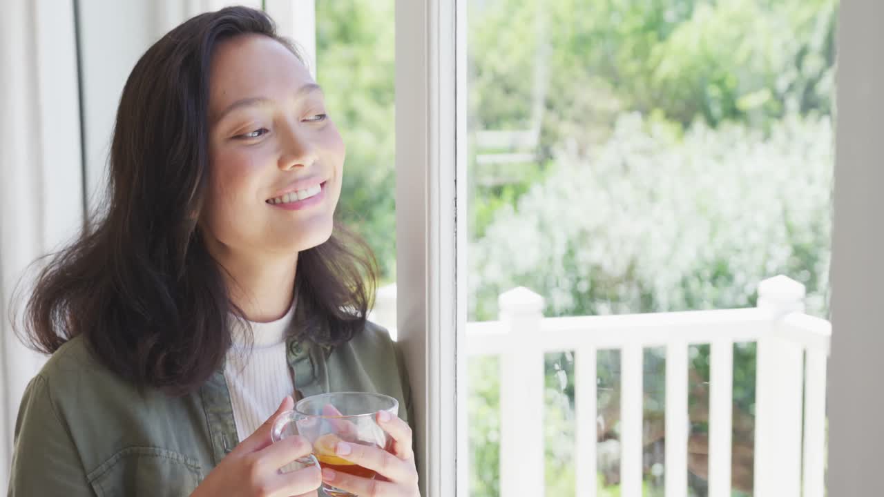 행복한 아시아 여성이 차를 마시고 슬로우 모션으로 창문을 통해보고 있습니다.