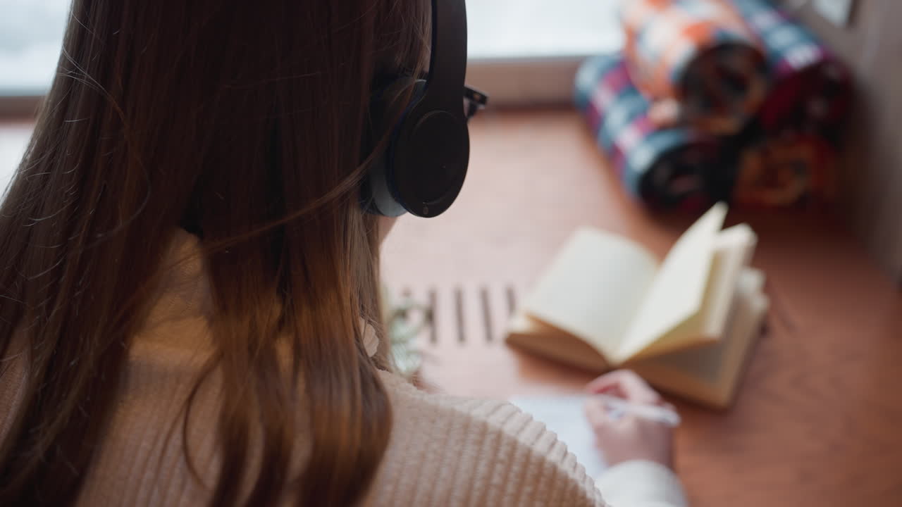 close up rear view of lady in headphone writing in notebook beside open book and folded cloth on wooden table with soft light entering through window