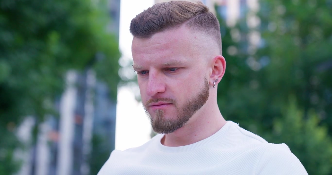 Giovane uomo concentrato con barba e piercing che guarda in basso all'aperto