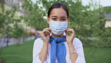 Alegre-Mujer-Asiática-Poniéndose-Una-Máscara-Médica-Al-Aire-Libre.-Vista-Frontal.-Ella-Sonríe-Después-Del-Bloqueo-Del-Coronavirus.-Pandemia-De-COVID-19