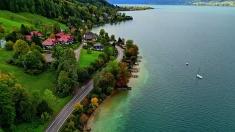 Vista-Aérea-Del-Attersee-En-Austria-Con-Casas-Junto-Al-Lago-Rodeadas-De-árboles-De-Colores-Otoñales-Y-Un-Velero