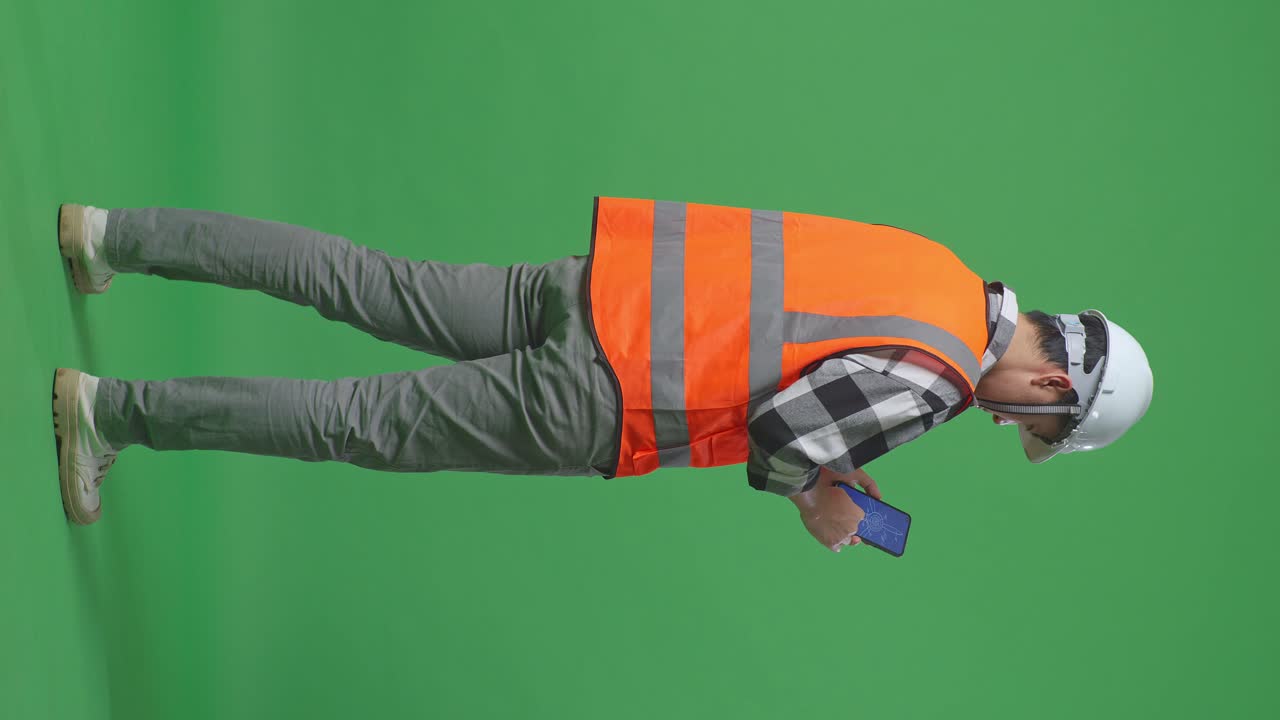 Full Body Back View Of Asian Male Engineer With Safety Helmet Looking At The Wind Turbine Blueprint On Smartphone While Standing In The Green Screen Background Studio