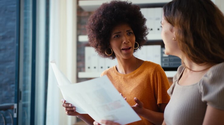 Two Young Female Business Colleagues With Documents Meeting Together In Office
