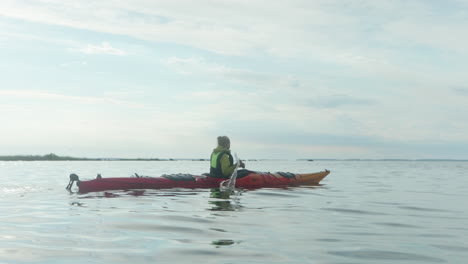 young blonde woman paddling in a red sea kayak on the open sea in finland, vaasa, archipelago, beautiful summer sunset atmosphere, wide shot