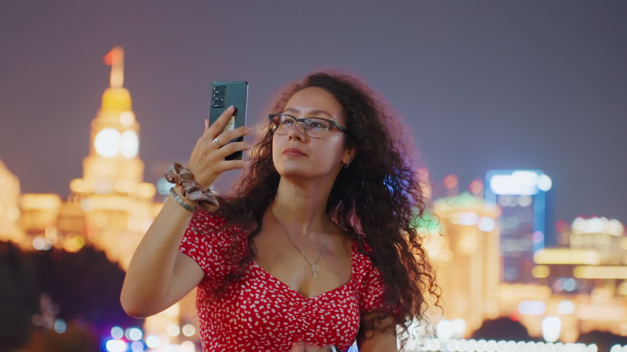 Svart kvinne tar selfie om natten, rød kjole, krøllete hår, briller, poserer mot en glødende byhorisont med bokeh-lys, leken, selvsikker turist fanger minner på telefonen, varm urban atmosfære