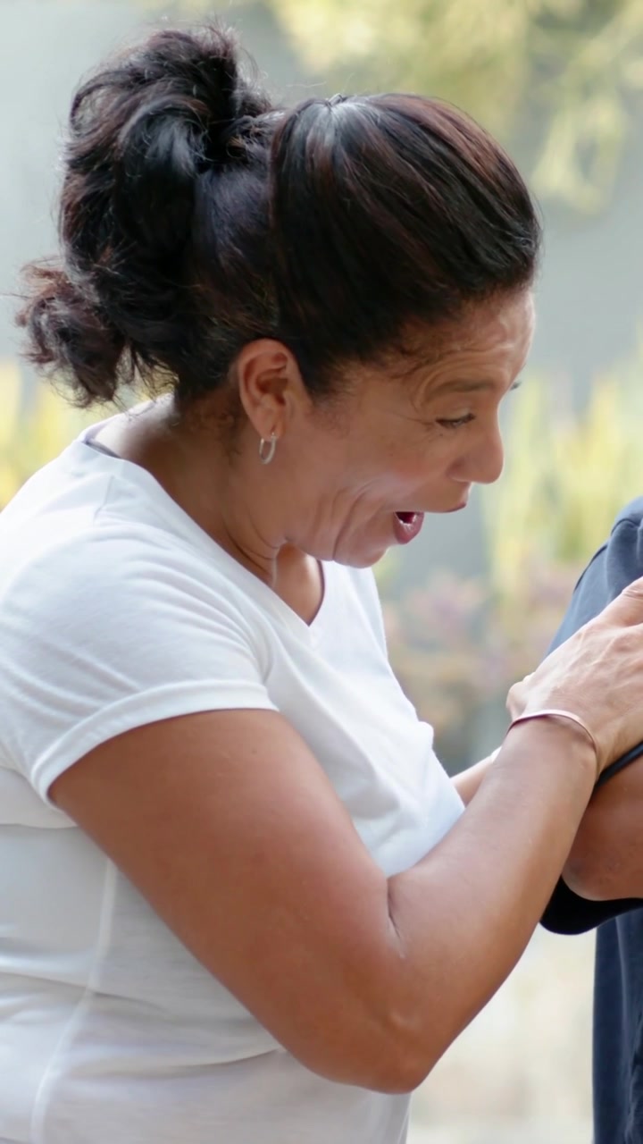 Video vertical: Mujer con camiseta blanca saludando, sonriendo, inclinándose, riendo, abrazando y moviéndose a la derecha en el patio.