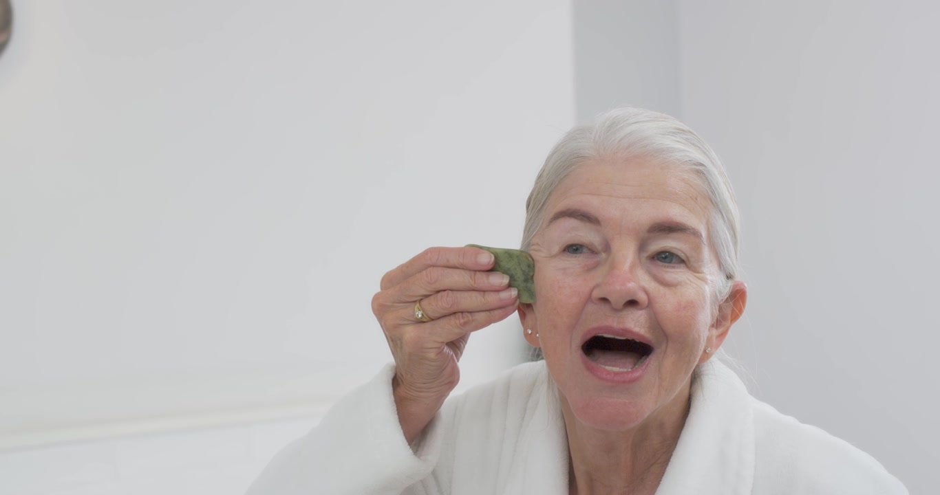 Donna anziana che apre la bocca con un gua sha verde, facendolo scorrere sulla mascella, sotto gli occhi o sulla fronte per il massaggio.
