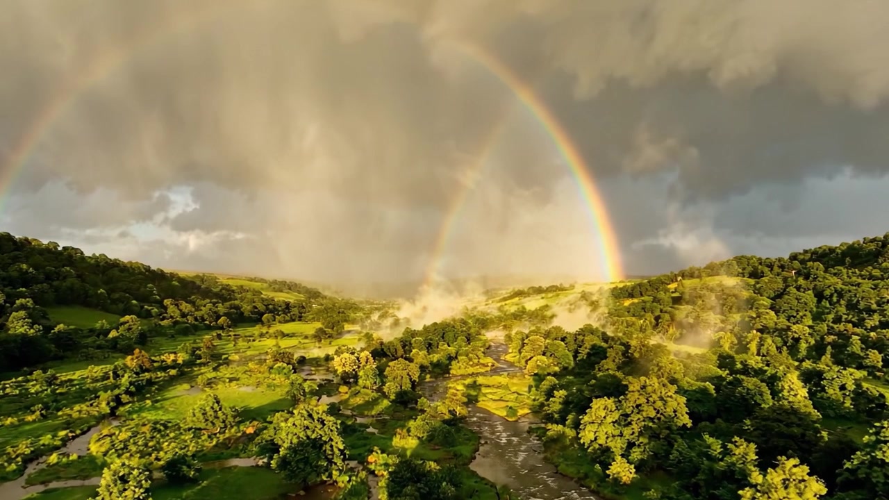 Pelangi Ganda yang Menakjubkan Melengkung di Atas Lembah Hijau Subur, Dikelilingi Kabut dan Pemandangan Awan Dramatis, Menangkap Keindahan Alam dalam Lanskap yang Tenang