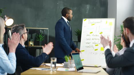 businessman standing at table and pointing at a blackboard while talking about a project with multiethnic business partners