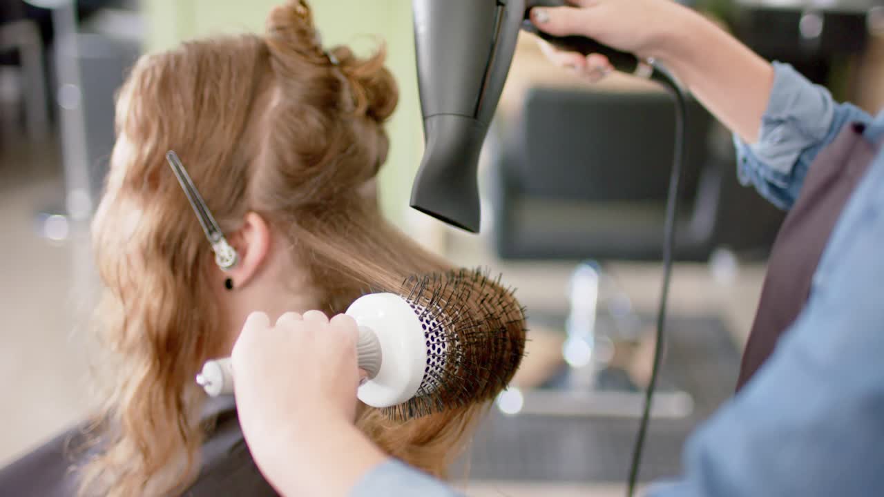 Caucasian female hairdresser styling client's long hair with hairdryer and brush, in slow motion