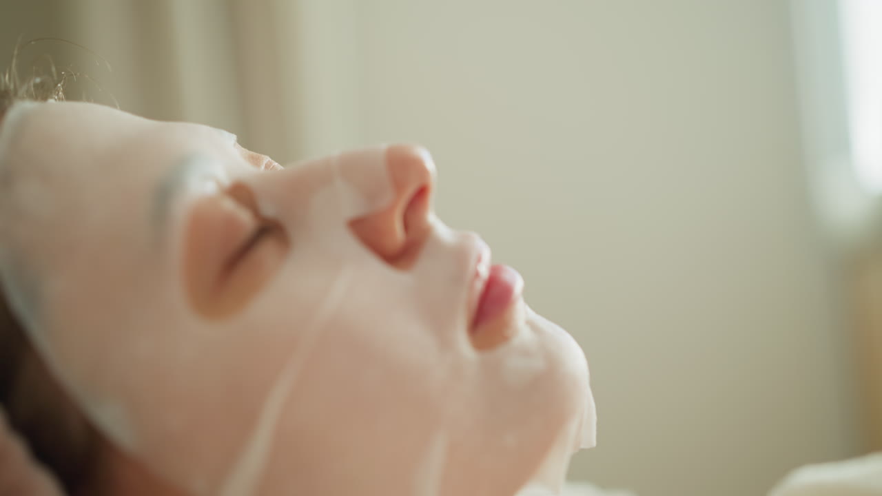 얼굴에 마스크를 쓴 여자가 침대에 누워서 음악을 듣고 있습니다. 매력적인 갈색 여성은 베개에 머리를 놓고 넓은 방에서 손을 움직입니다.