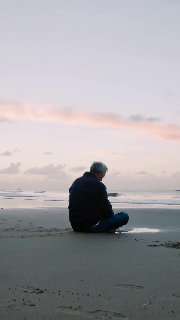 Vertical video: Sitting older man shifting, thinking at sunrise by pool on beach in jacket, jeans