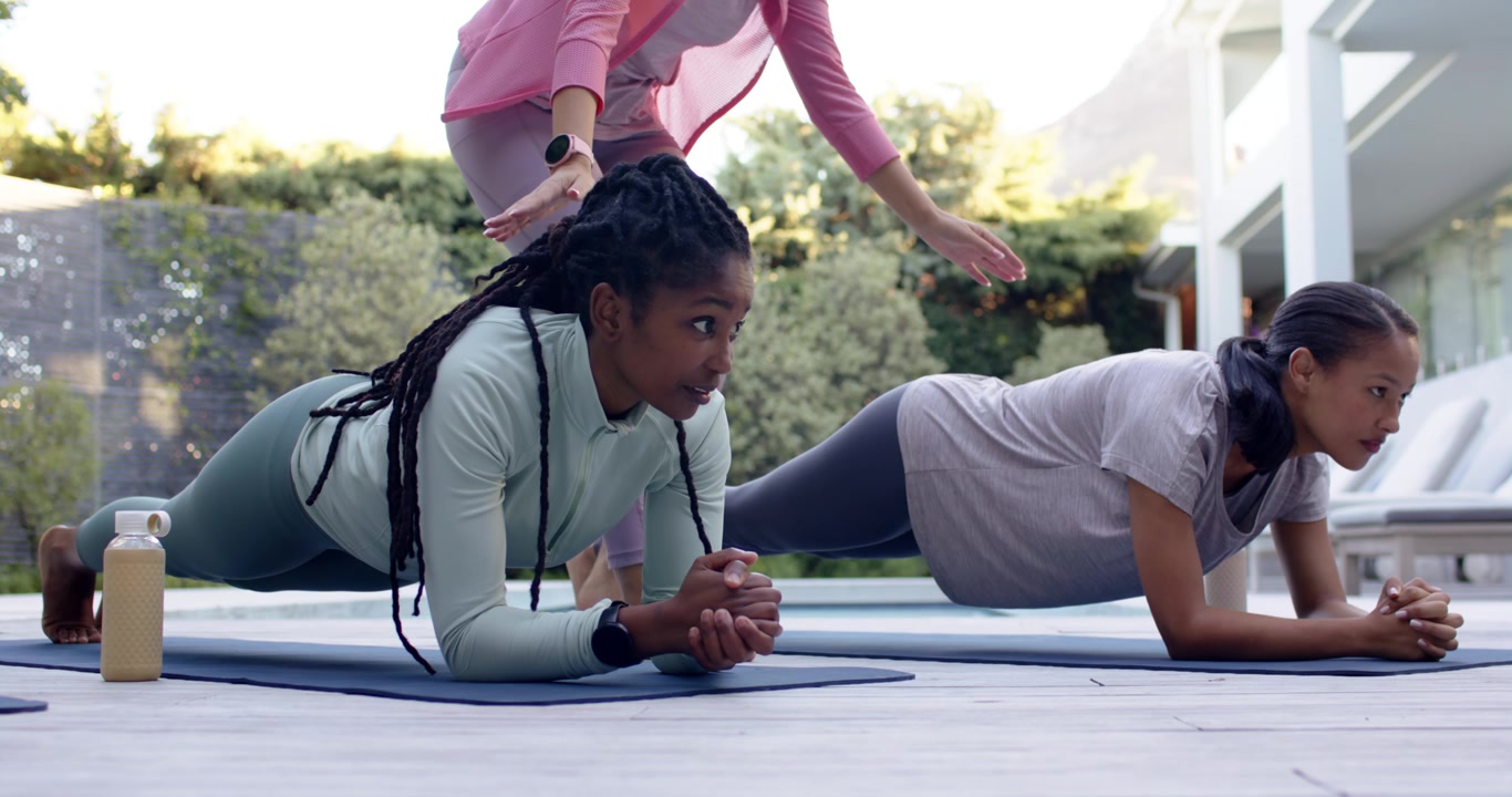 Diverse women holding forearm plank on yoga mats on patio as trainer walking adjusting alignment
