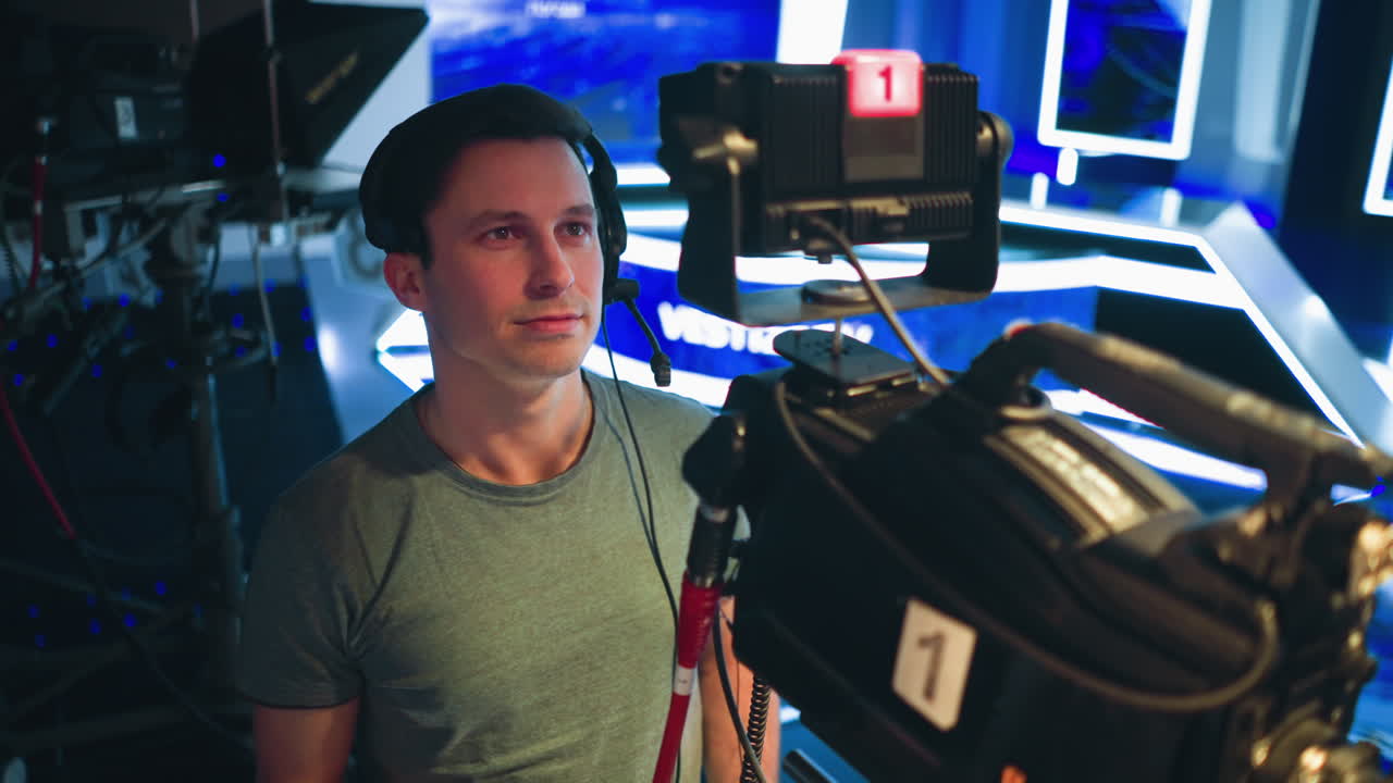 Male cameraman wearing headset operates professional studio camera during live television broadcast, standing in high-tech set with illuminated background screens and control equipment visible around filming space