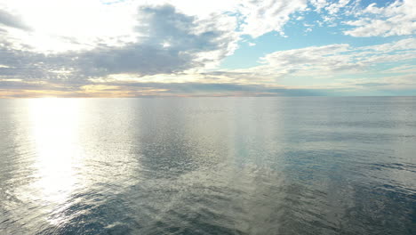 aerial: flying backwards with calm and peaceful sea with reflection of sky and cloudy sky in the background