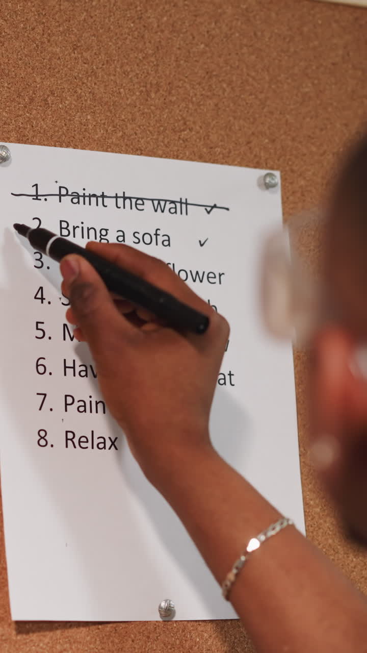 Black guy crosses out accomplished to-do list item on cork board near grunge wall closeup. African-American worker with plan of flat renovation process