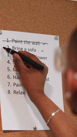 Black guy crosses out accomplished to-do list item on cork board near grunge wall closeup. African-American worker with plan of flat renovation process