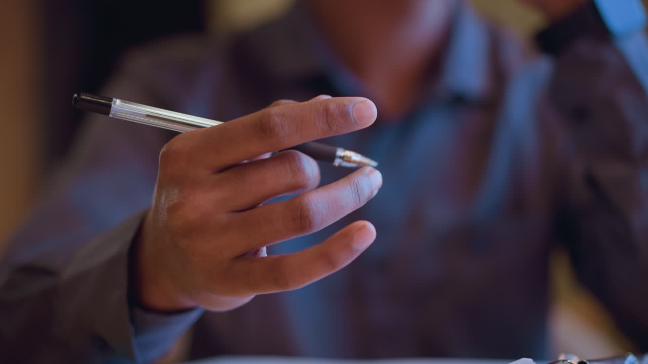 Close-up of man holding pen between fingers while sitting thoughtfully with chin resting on hand, expressing quiet contemplation, decision making, or focused attention in relaxed indoor environment