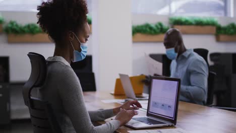 Diverse-male-and-female-business-colleagues-in-face-masks-using-laptops-in-office