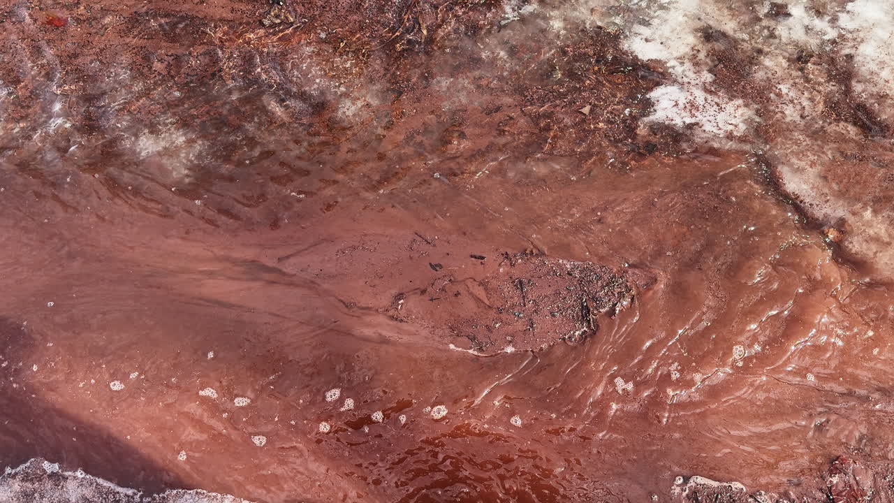 Acqua di disgelo limpida scorre su terreno rosso che si sta scongelando e ghiaccio persistente, rivelando ricche texture naturali in una vivida scena di fine inverno dove la primavera inizia a prendere piede