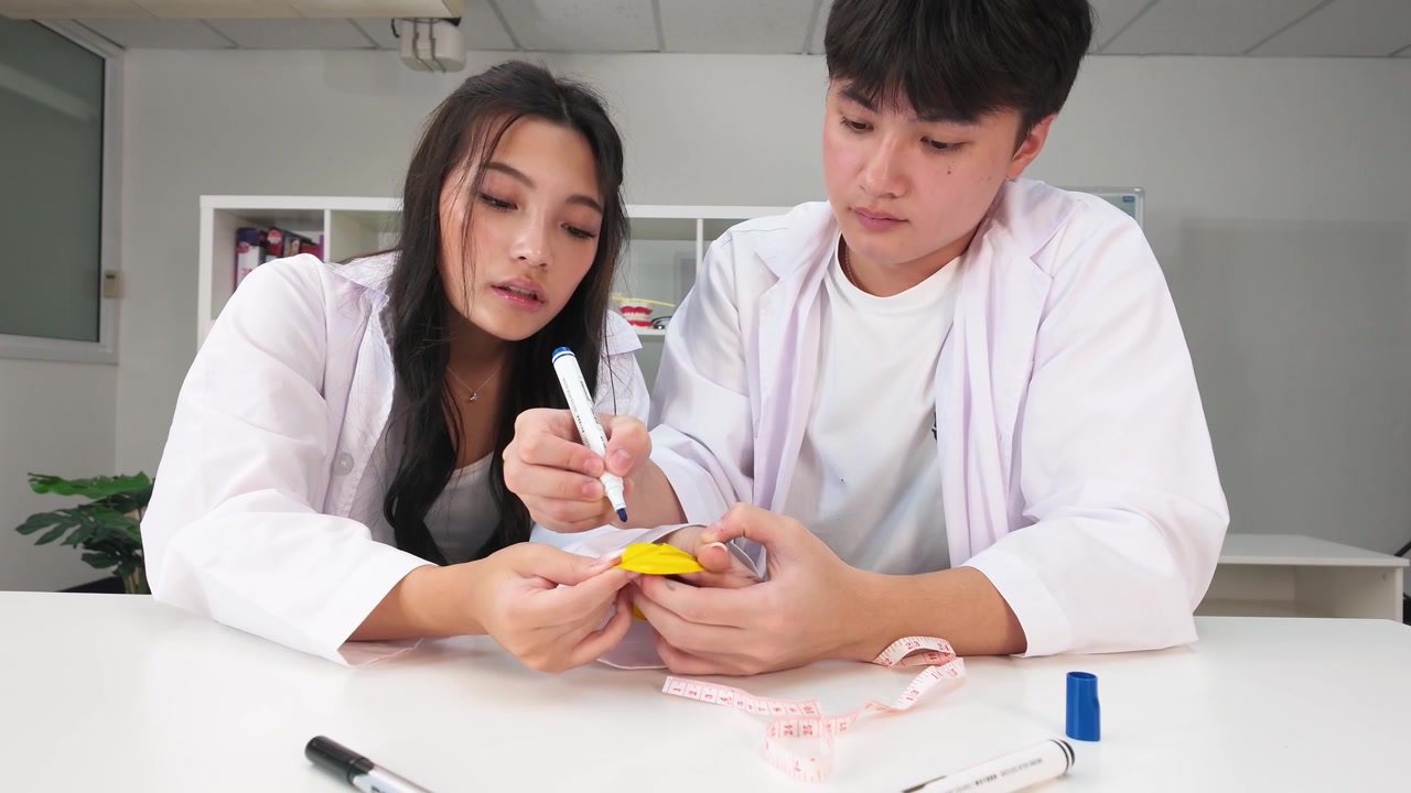 教室で白衣を着た2人のアジア人学生が科学実験に協力している。