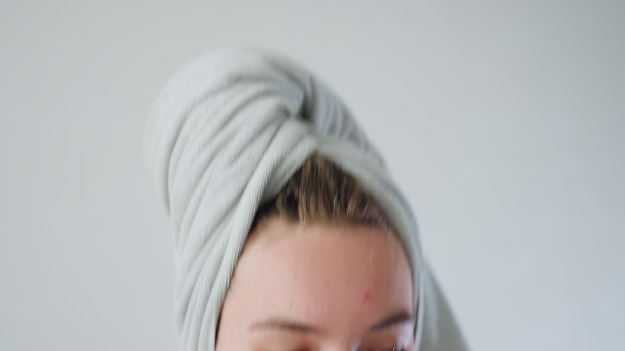 Close profile view of young woman with towel wrapped around head and visible acne blemishes sitting on sofa filing nails with nail file in bright living room with sheer curtains and chandelier