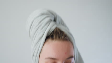 close profile view of young woman with towel wrapped around head and visible acne blemishes sitting on sofa filing nails with nail file in bright living room with sheer curtains and chandelier