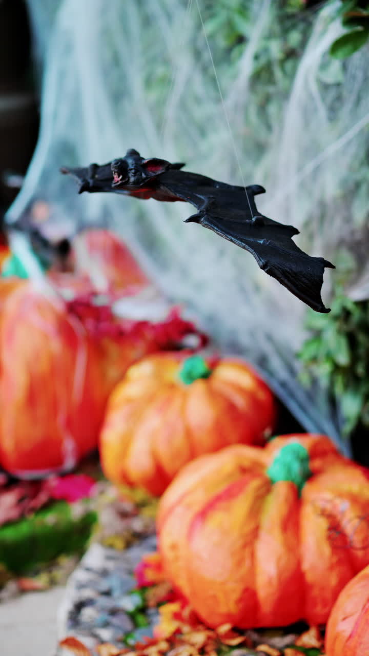 A bat figurine suspended above halloween decorations of pumpkins and spider webs. Vertical