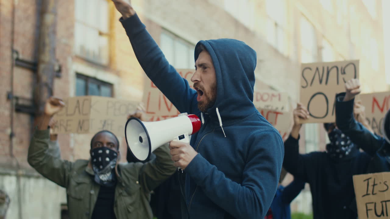 Caucasian man with hoodie and arms up yelling on a loudspeaker in a protest with multiethnic group of people in the street