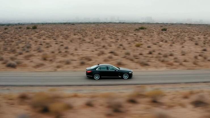 Side view shows black sedan moving along straight desert highway beside scrub and sand, with distant hazy skyline, chrome trim, tinted windows, soft overcast light, spacious travel mood