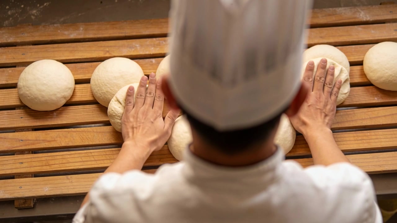 Noticing row of dough, baker wearing chef's uniform shaping rounds on wooden bench for proofing