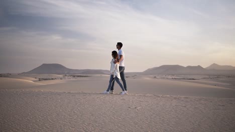 Imágenes-En-Cámara-Lenta-De-Una-Pareja-Abrazada-Caminando-En-Una-Relación-Romántica-Bajo-El-Sol-Y-El-Cielo-Azul-En-El-Desierto.-Dos-Jóvenes-Amantes-Caminando-Por-La-Arena-Del-Desierto-Con-Ropa-Informal.-Perfecta-Escena-Borrosa-De-Montañas-En-El-Fondo