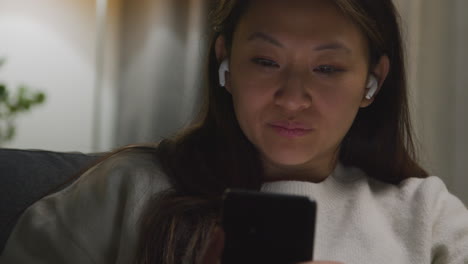 close up of woman spending evening at home on sofa wearing wireless earbuds and streaming music or podcast from mobile phone 1