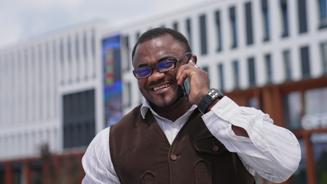 Portrait of positive american man with a phone. African american businessman in stylish glasses talking the phone and smiles on camera in the city.
